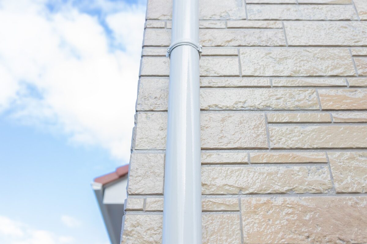 外壁の角部分を接写した写真。ベージュ系の石目調サイディングに、白い縦樋（雨どい）が中央に通っている