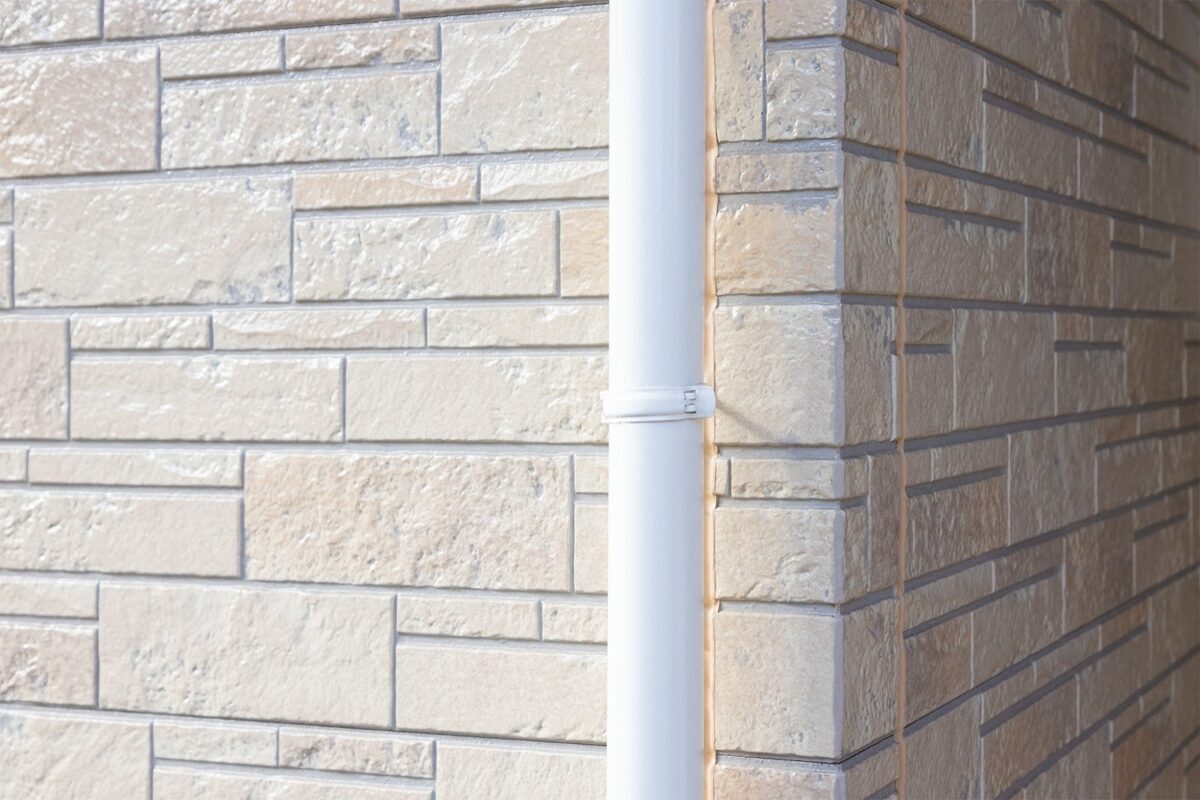 ベージュ系の石目調サイディングの上に、白い縦樋（雨どい）が固定されていて、角のコーキング（シーリング）部分が見える