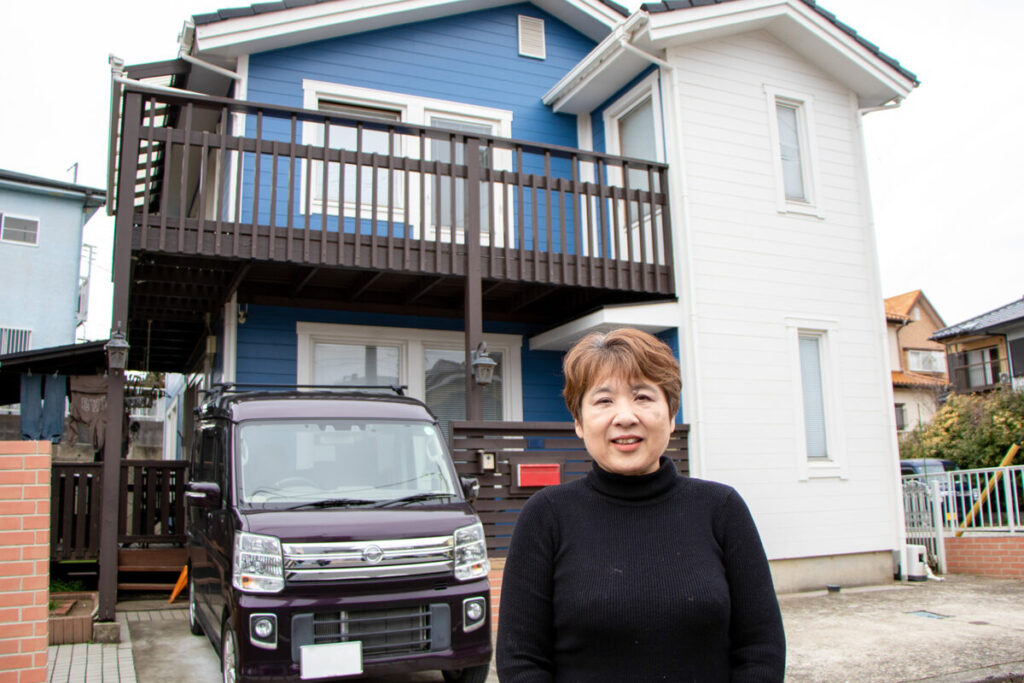 青と白のツートンカラーの北欧住宅の前に立つH様。2階には木製のバルコニーがある。